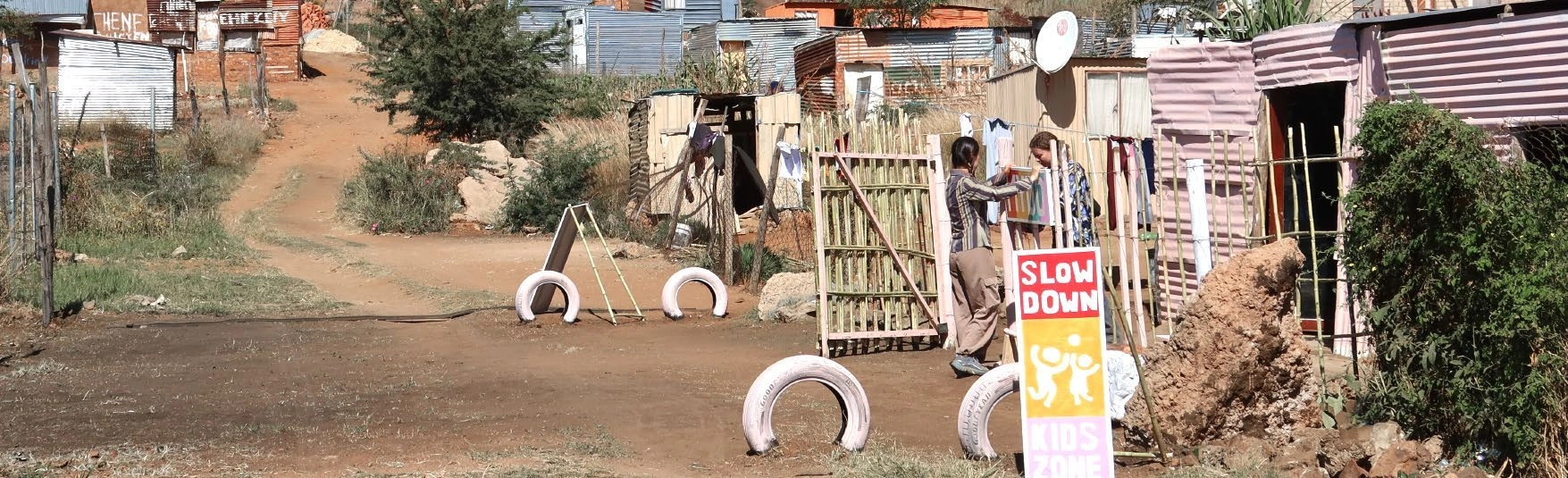 Built roadside children’s zone with painted tyres, a reed fence, and a Slow Down Kids Zone sign.
